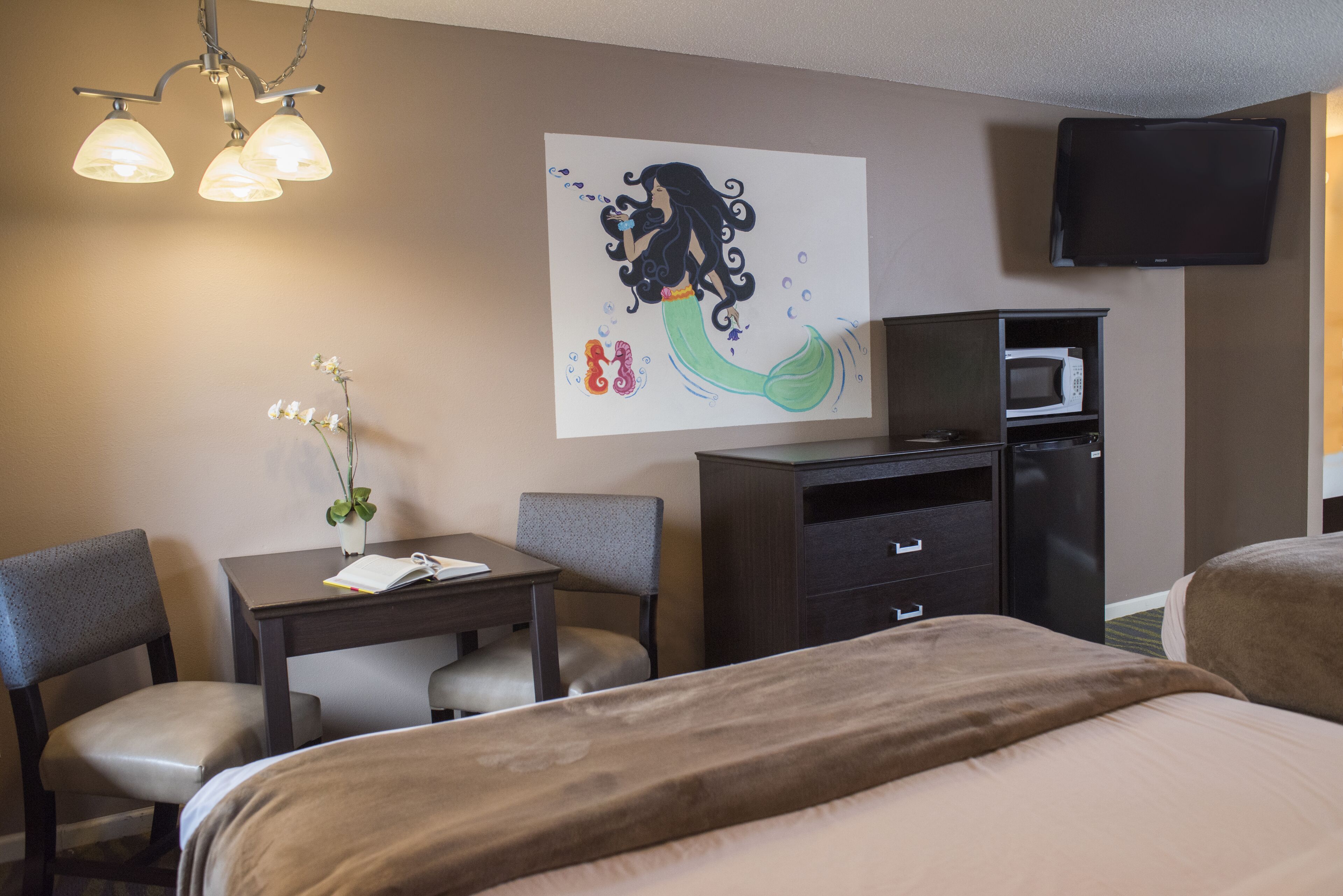 A hotel room with a bed, a desk with a chair, a small table with a plant, a flat-screen TV mounted on the wall, and a microwave in the kitchen area.