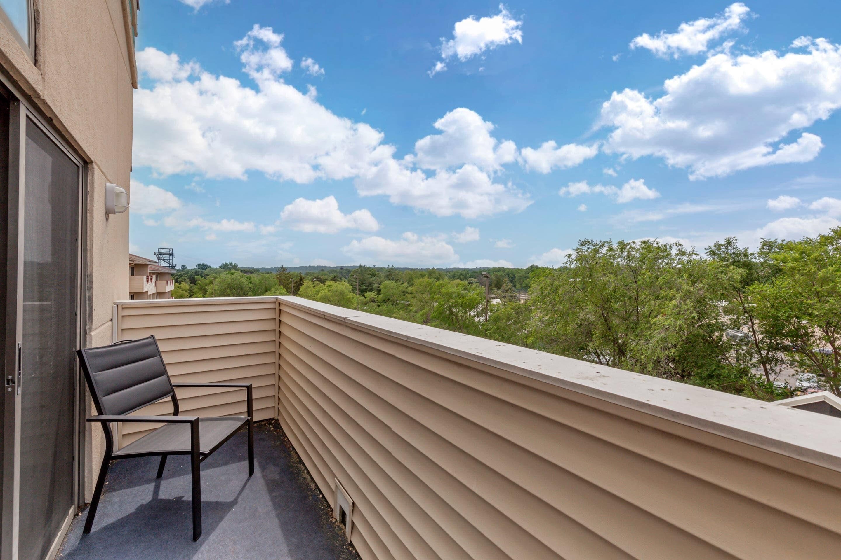 A balcony with a chair, beige siding, and a view of greenery and buildings under a partly cloudy sky.