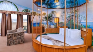 Una cama redonda de madera con dosel, un sillón estampado y un mural de una playa tropical.