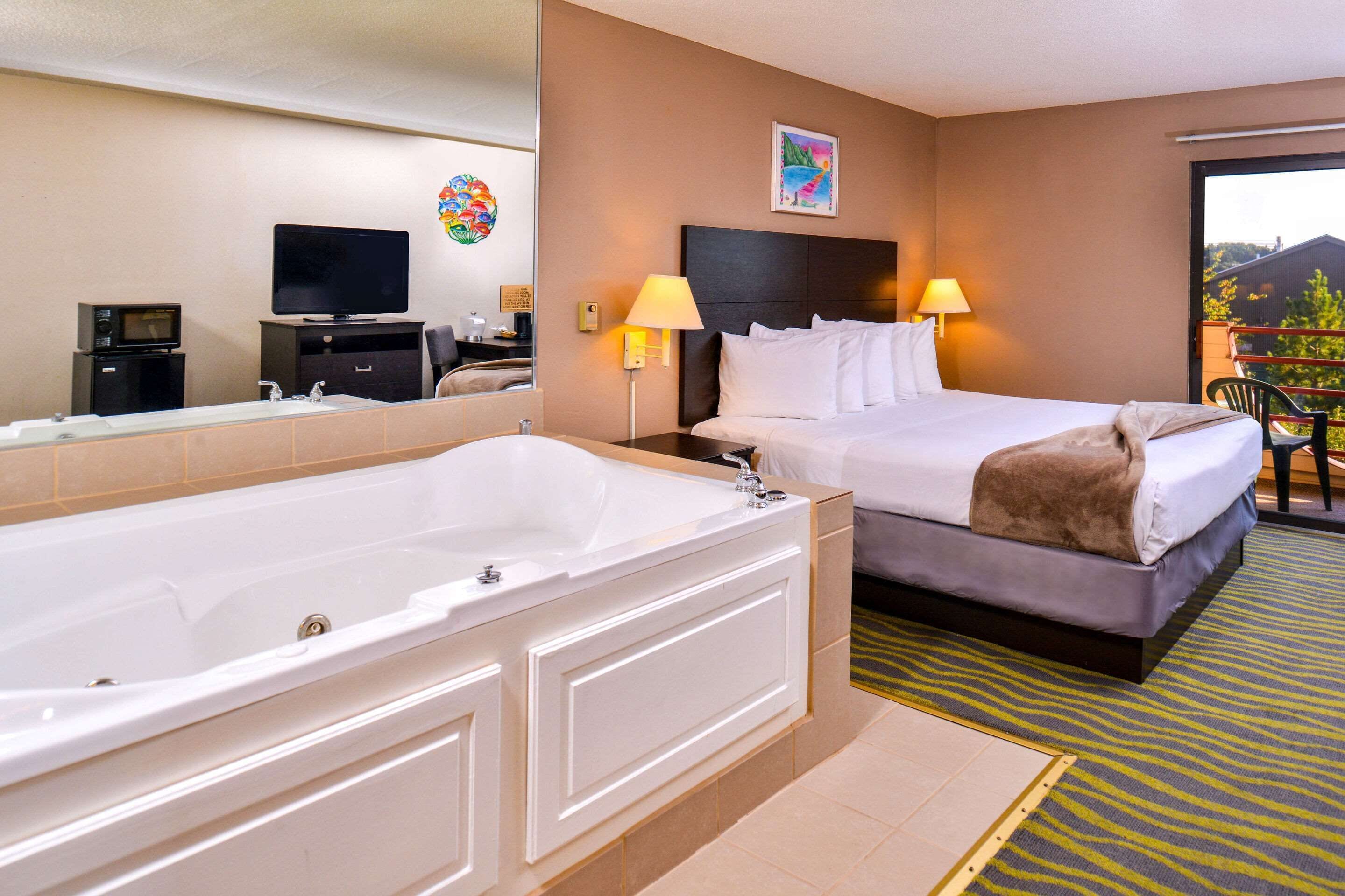 Habitación de hotel con una bañera blanca grande, cama con ropa de cama blanca, televisor de pantalla plana, escritorio con computadora y balcón con vistas.