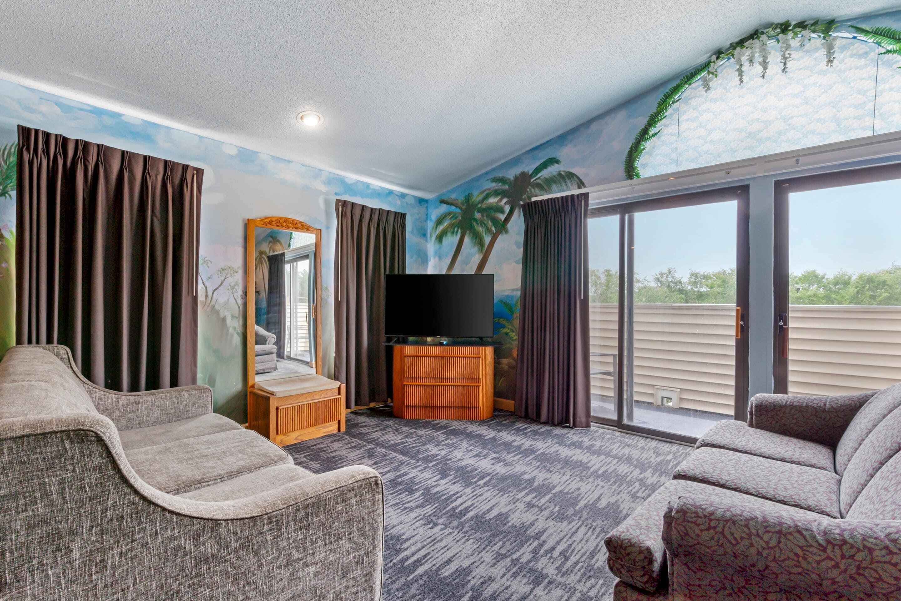 A hotel room with a large window, a flat-screen TV on a wooden stand, a sofa, and a carpeted floor.