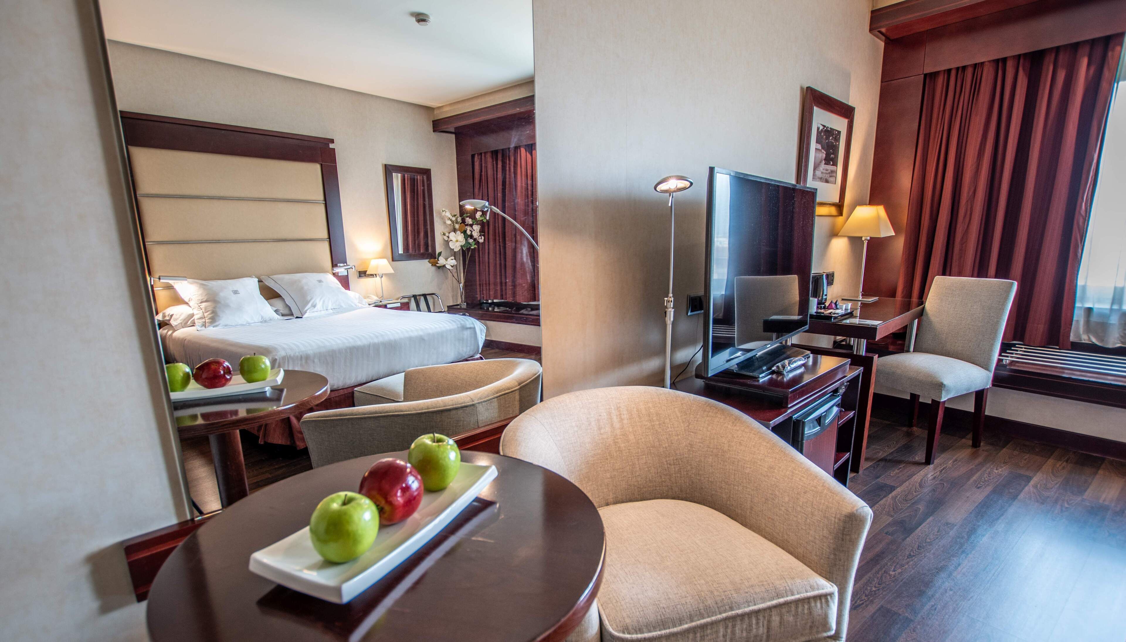 A hotel room with a bed, a desk with a TV, a chair, and a table with apples.