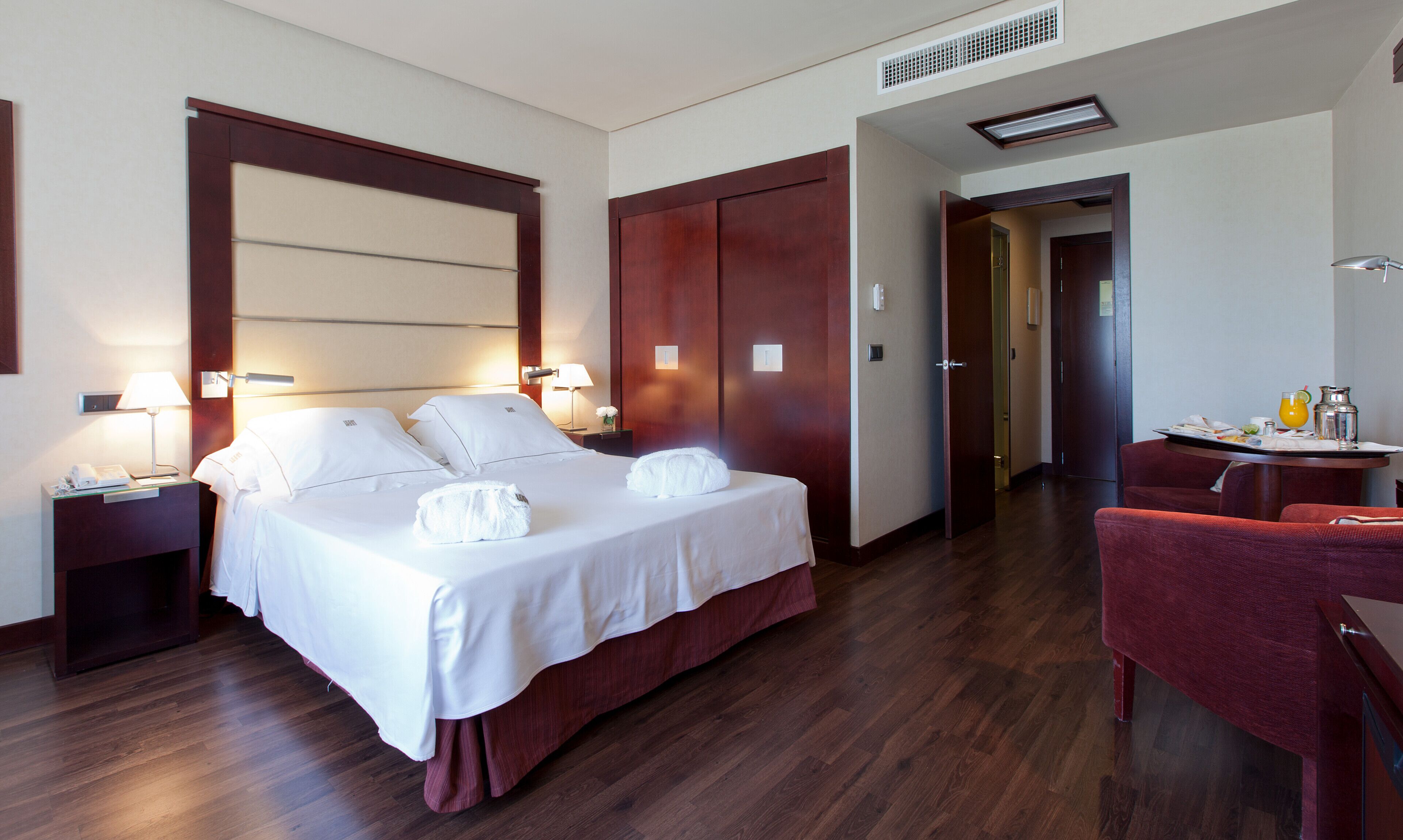 A hotel room with a large bed, bedside lamps, a desk with a chair, and a door leading to another room.