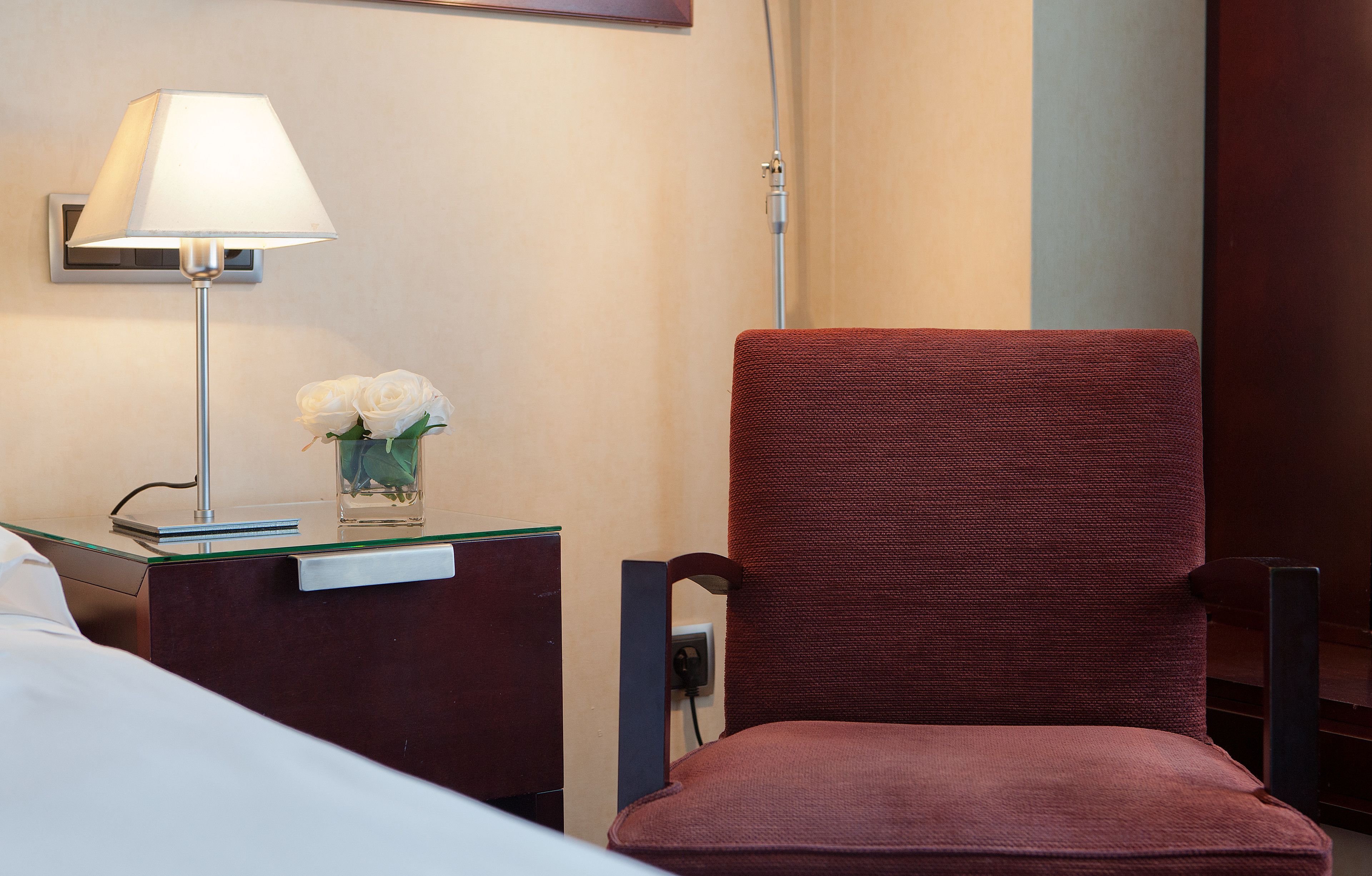 A hotel room with a red upholstered chair, a glass table with a lamp and a vase of white flowers, and a bed.