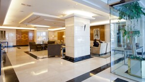 Un hall d’entrée spacieux au design moderne, comprenant un grand espace salon avec des canapés et une table basse, un comptoir d’accueil et un mur de verre agrémenté d’une plante.