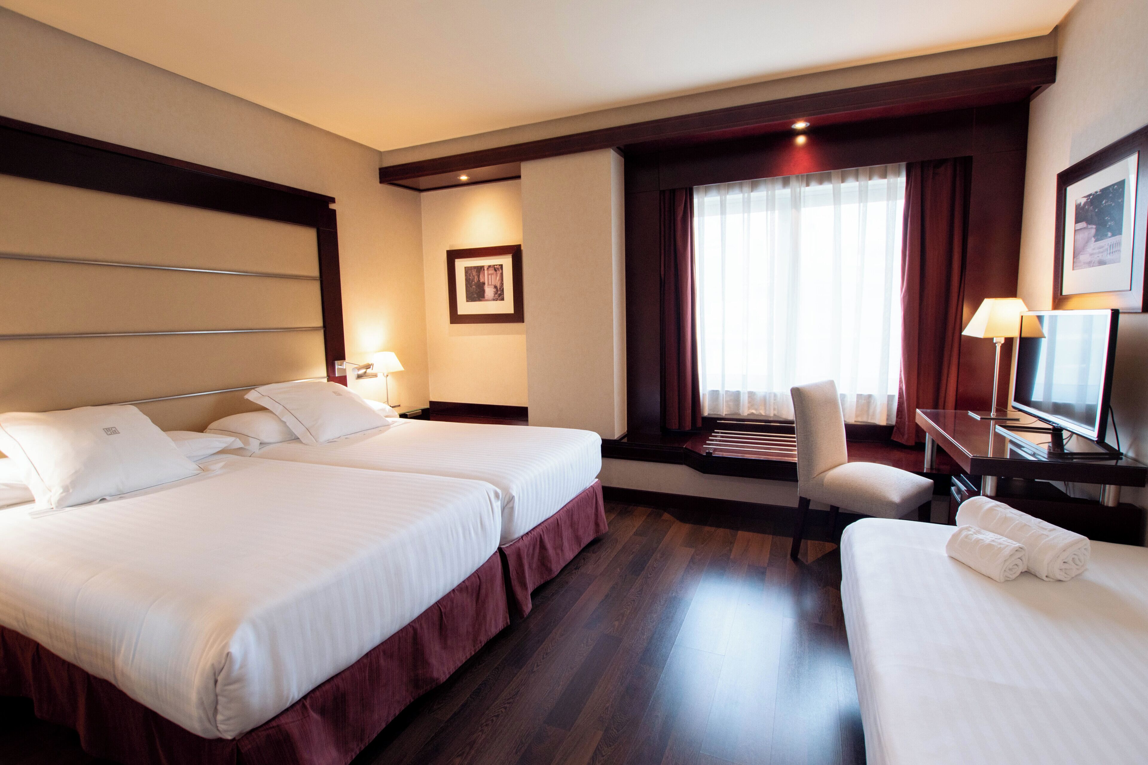A hotel room with two beds, a desk, a chair, a TV, and a window with curtains.