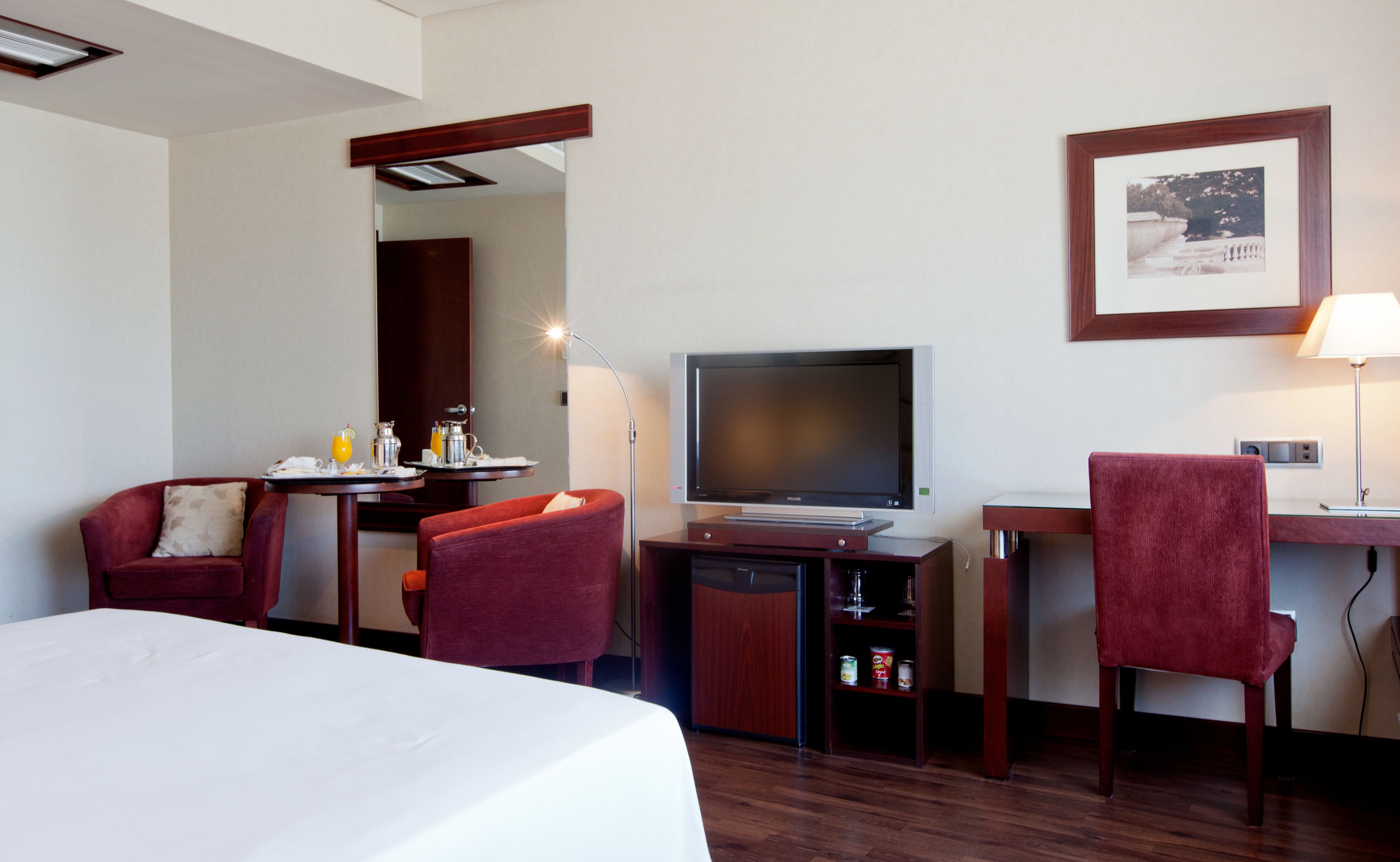Habitación de hotel con cama, escritorio con televisor, dos sillas, una pequeña mesa con productos de desayuno y un cuadro en la pared.
