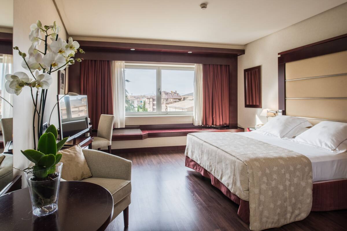 A hotel room with a bed, a TV, a chair, a desk, and a window with curtains.