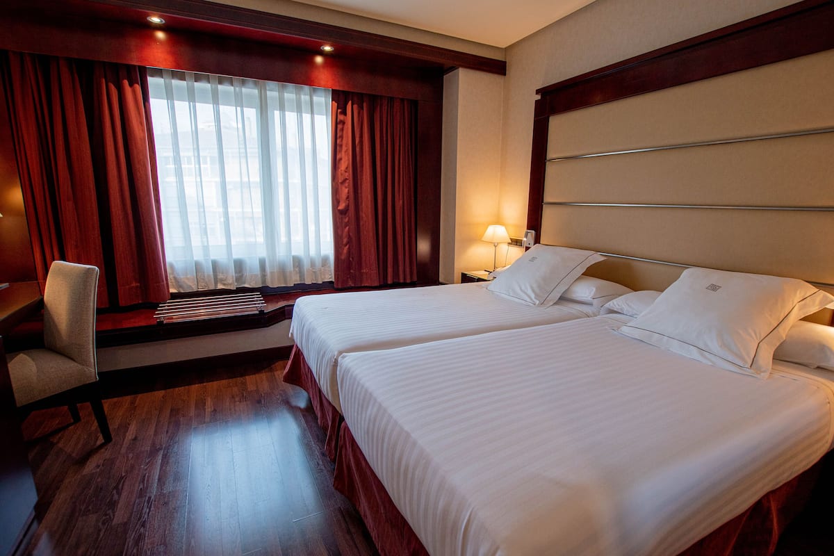 A hotel room with two beds, a chair, a window with curtains, and a nightstand with a lamp.