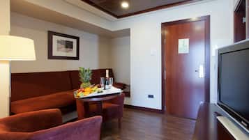 A hotel room with a wooden floor, a brown sofa, a small table with fruit and a bottle, a TV, and a door with a sign.