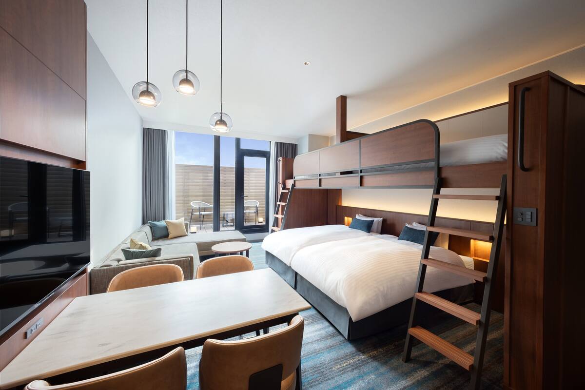 Una habitación de hotel moderna con literas, una zona de comedor con una mesa y sillas, un televisor y un amplio ventanal con vistas al exterior.
