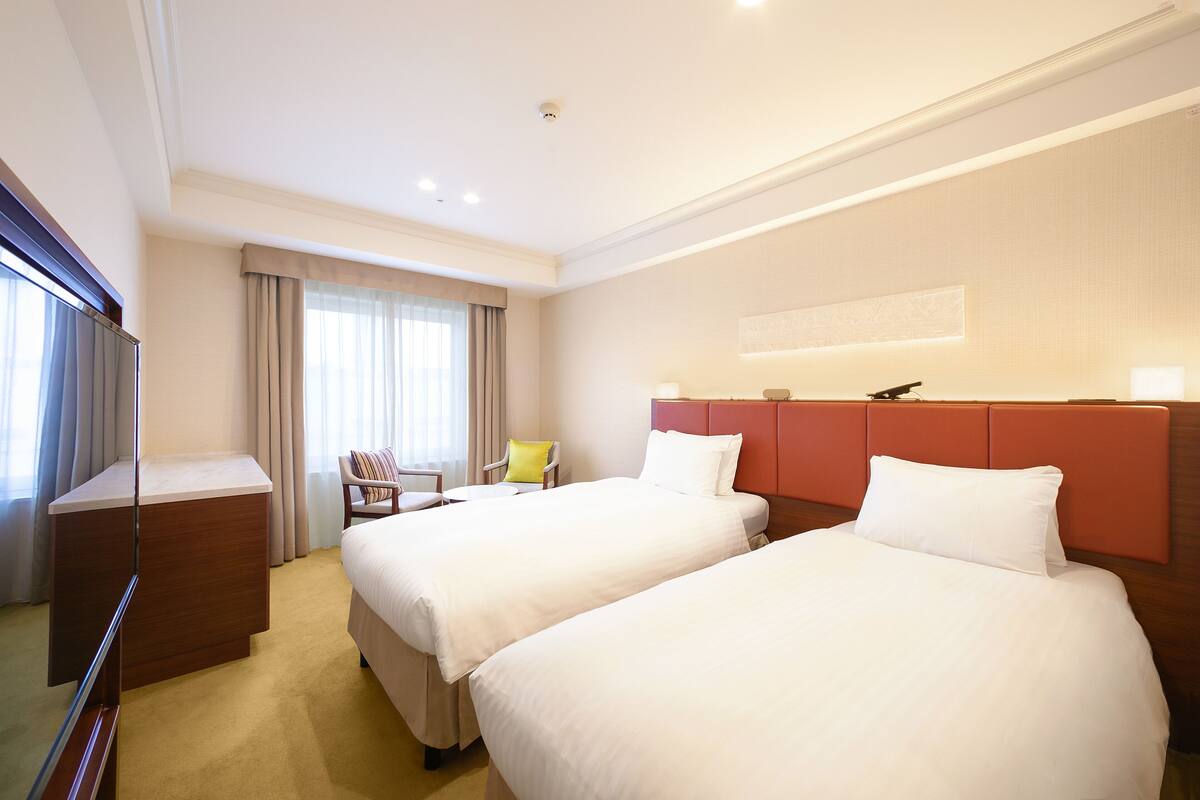 Habitación de hotel con dos camas, un escritorio, una silla y un ventanal con cortinas.