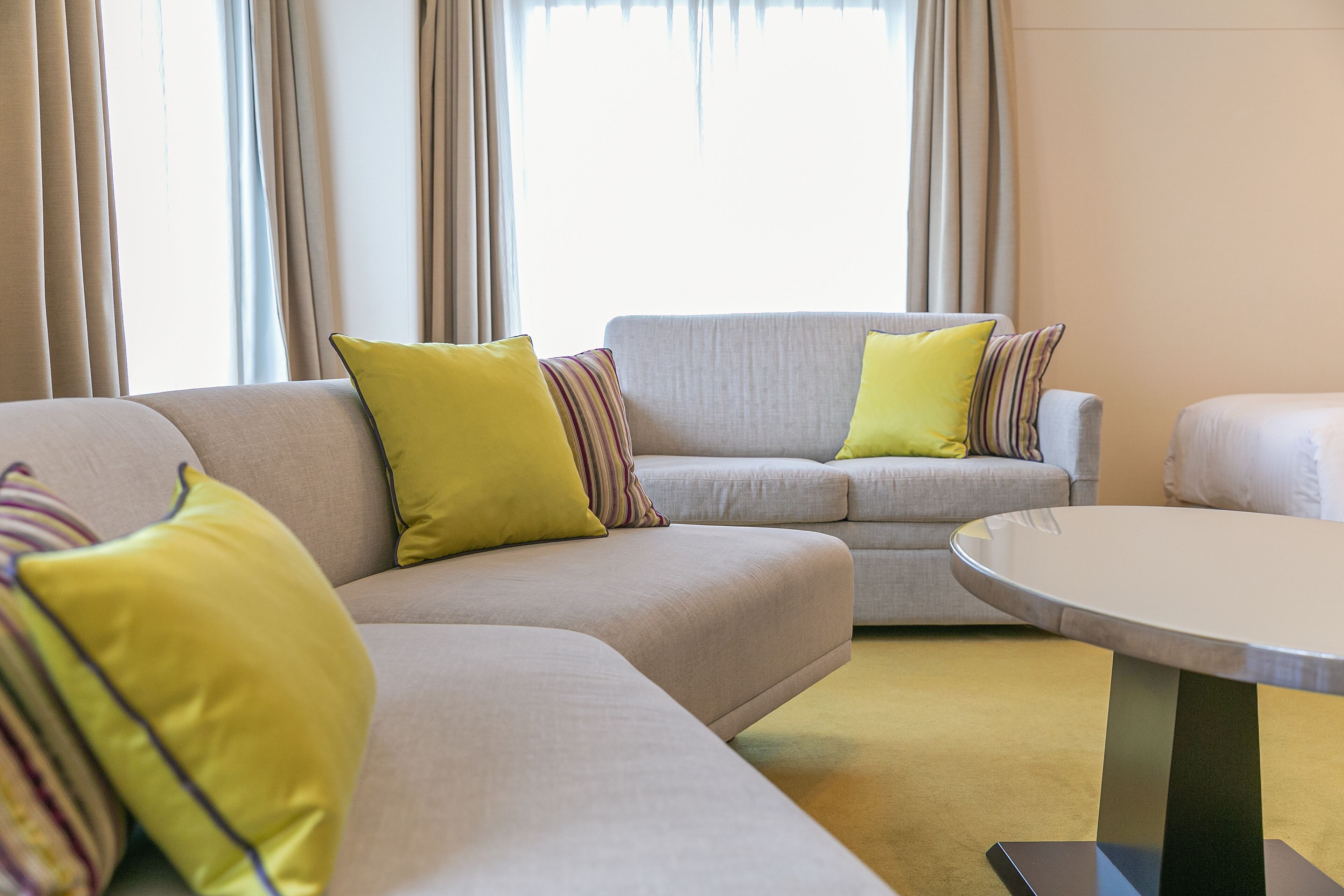 A modern living room with a beige sofa and yellow pillows, a round table, and large windows with curtains.