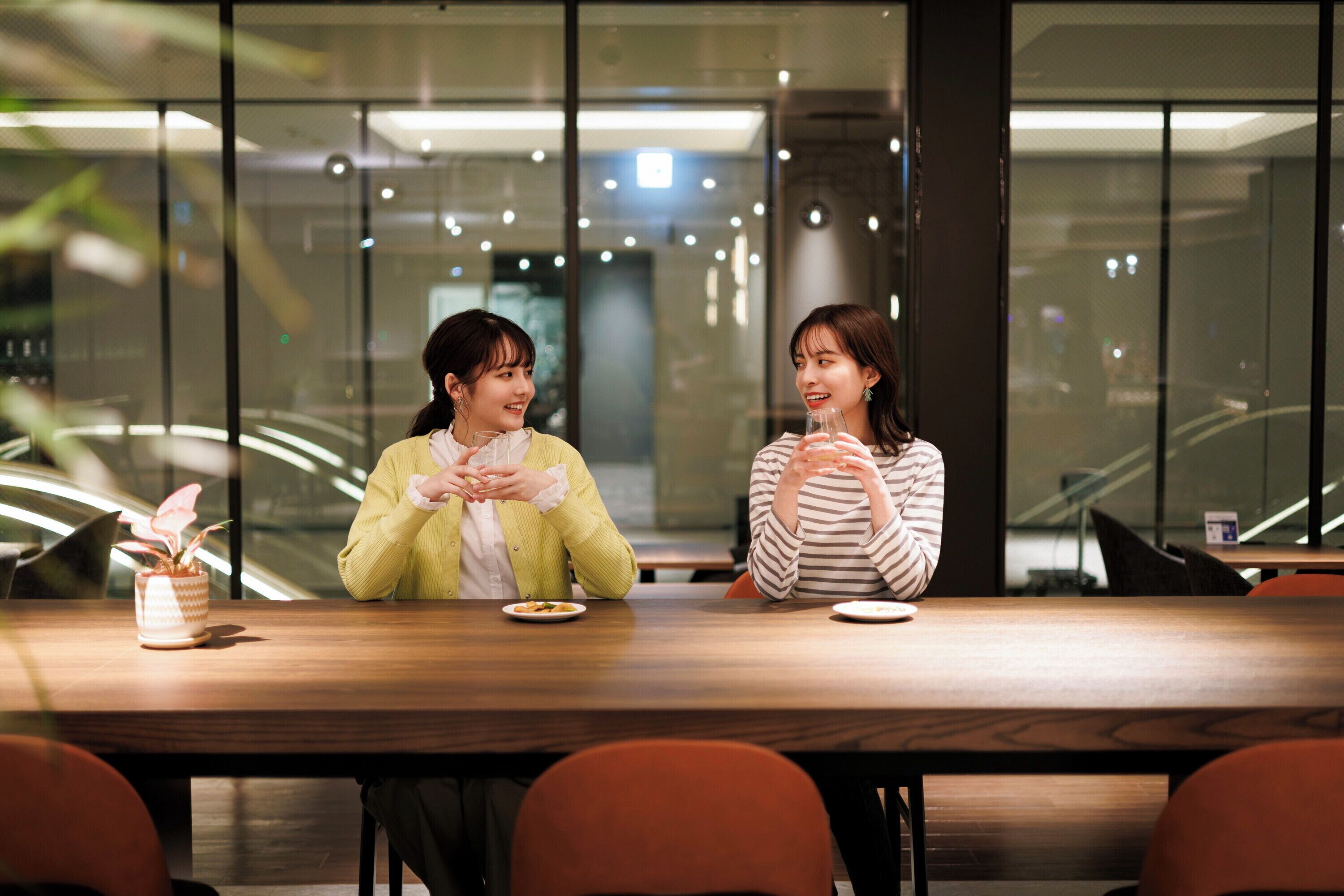 Two people sitting at a wooden table in a modern indoor setting with glass walls and ceiling lights.