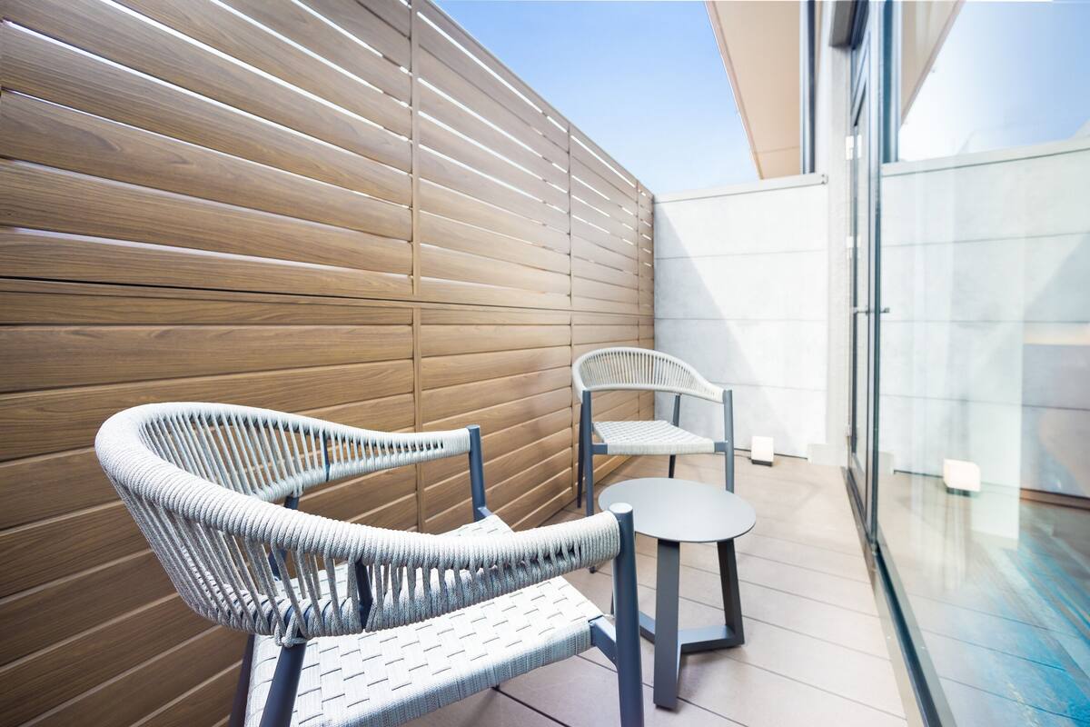 Un área de estar exterior moderna con sillas de mimbre, una pequeña mesa, pared de listones de madera y un separador de vidrio.
