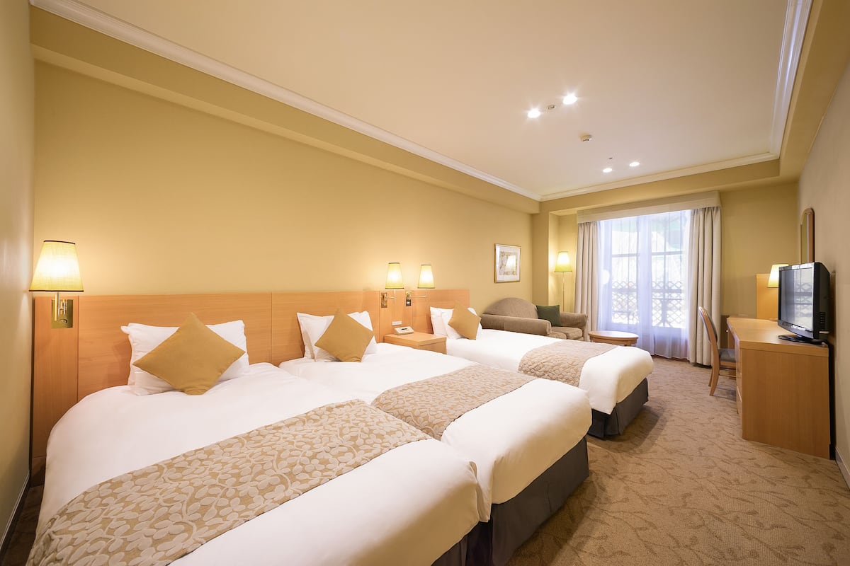 Habitación de hotel con tres camas, un escritorio, televisor y un amplio ventanal con cortinas.