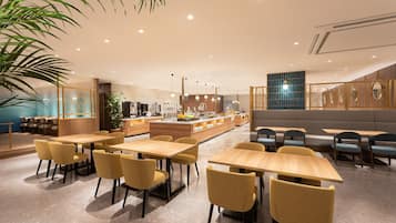 A modern dining area with wooden tables and yellow chairs, a kitchen in the background, and a decorative plant on the left.