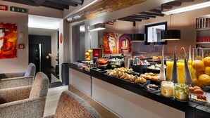 Un bufé de desayuno con diversos alimentos y bebidas, una zona de estar con sillones tapizados, y una decoración moderna con vigas a la vista y obras de arte en las paredes.