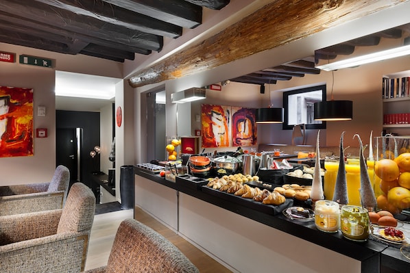 Un buffet de petit-déjeuner proposant une variété d’aliments et de boissons, un espace détente avec des fauteuils confortables, et un décor intérieur moderne avec des poutres apparentes et des œuvres d’art aux murs.