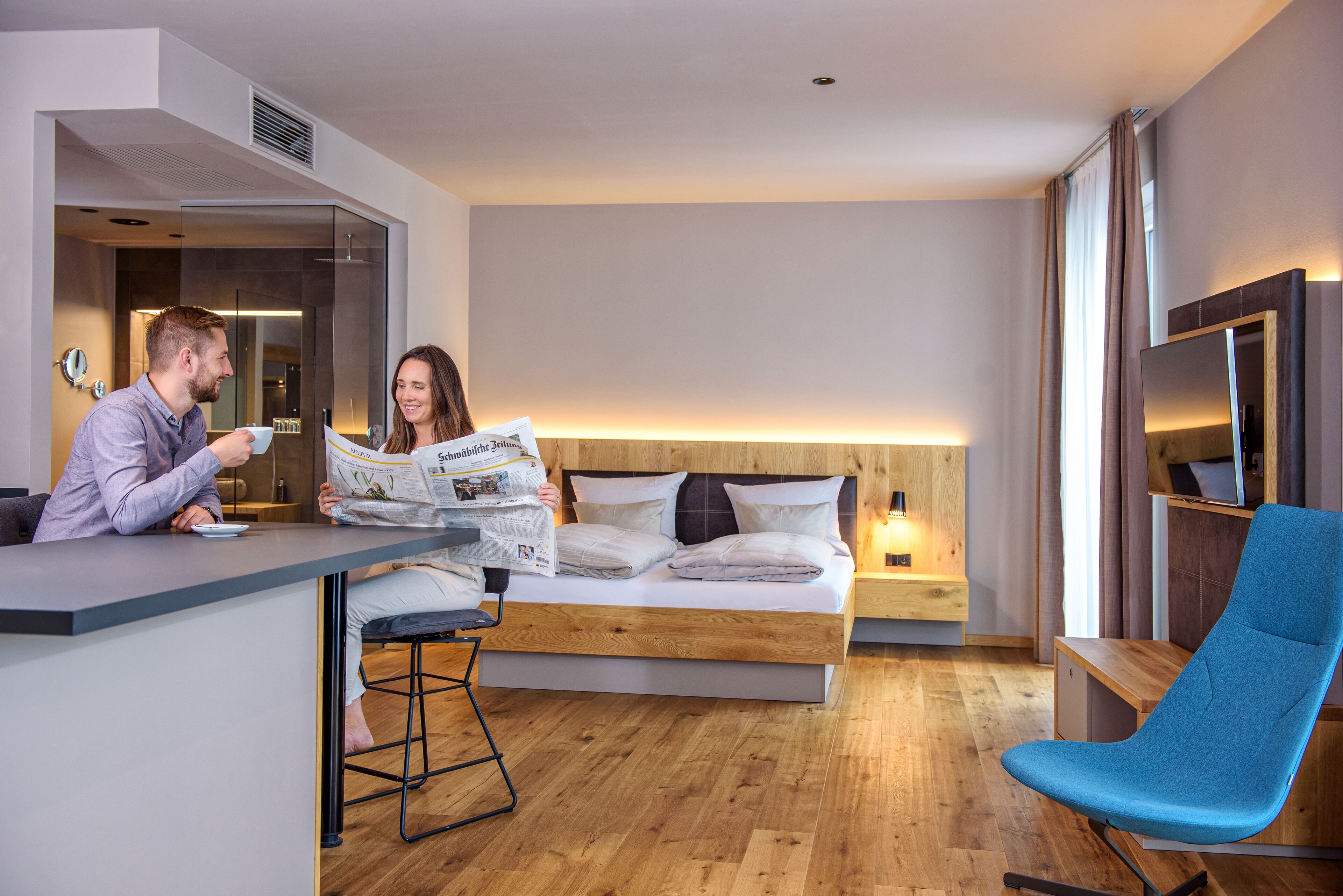 A modern hotel room with a wooden bed, a blue chair, and a breakfast bar.