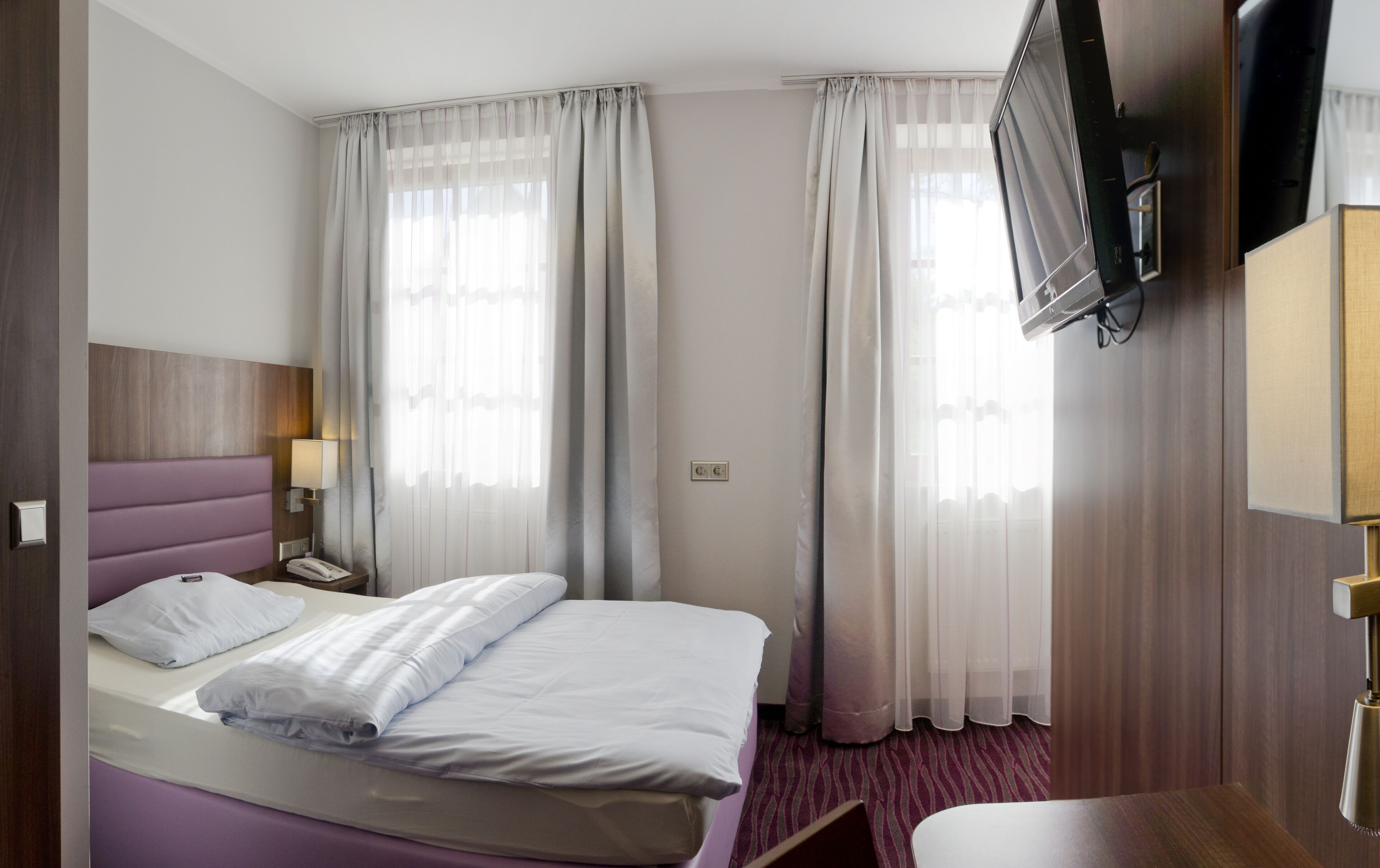 Habitación de hotel con cama, mesita de noche con lámpara, televisión montada en la pared y ventana con cortinas.