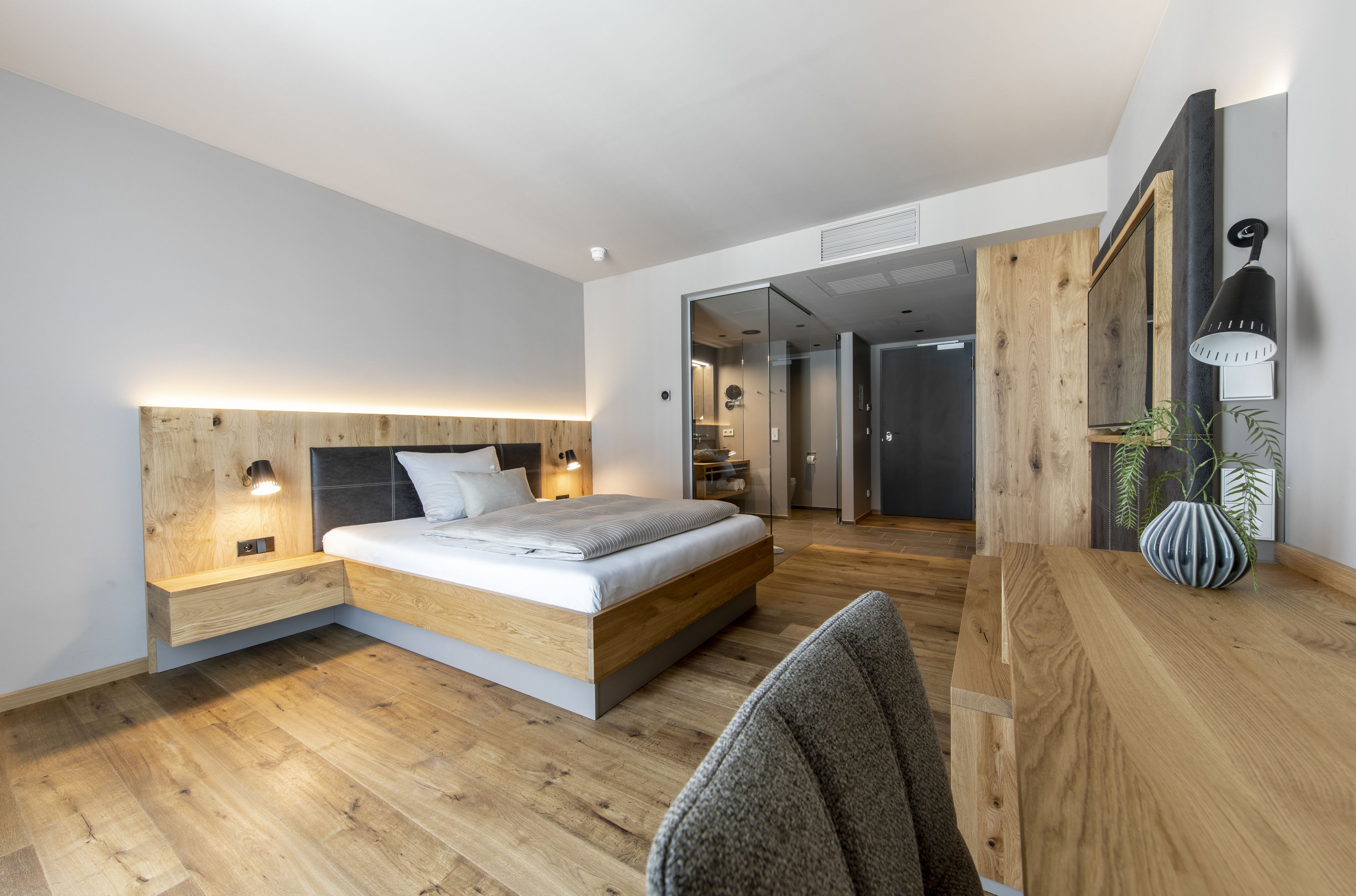 Une chambre à coucher moderne avec une tête de lit en bois, un lit recouvert de linge blanc, une table de chevet avec une lampe et un bureau en bois agrémenté d’un vase.