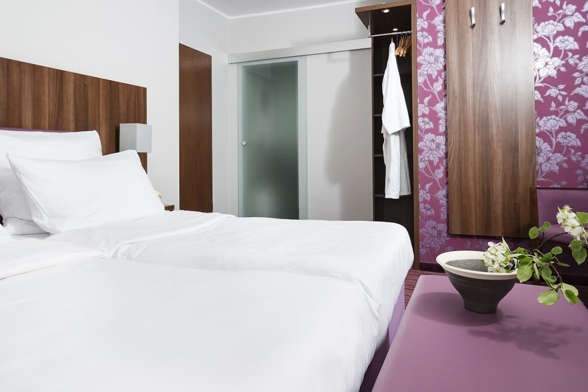 Une chambre d’hôtel avec deux lits, une armoire, un miroir et une petite table sur laquelle se trouve un vase avec des fleurs.