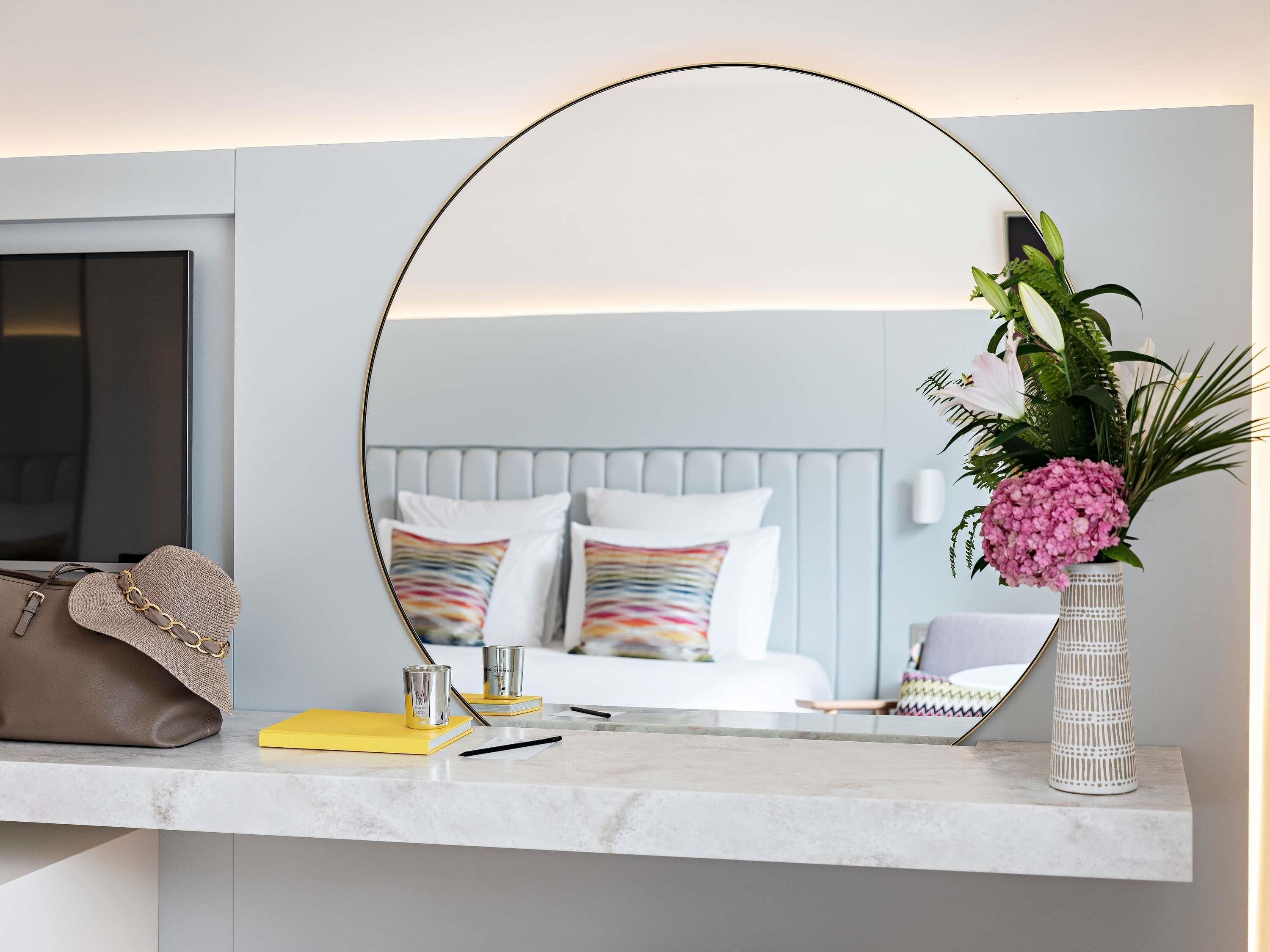 A hotel room with a bed, a bedside table with a mirror, a vase with flowers, and a suitcase on the floor.