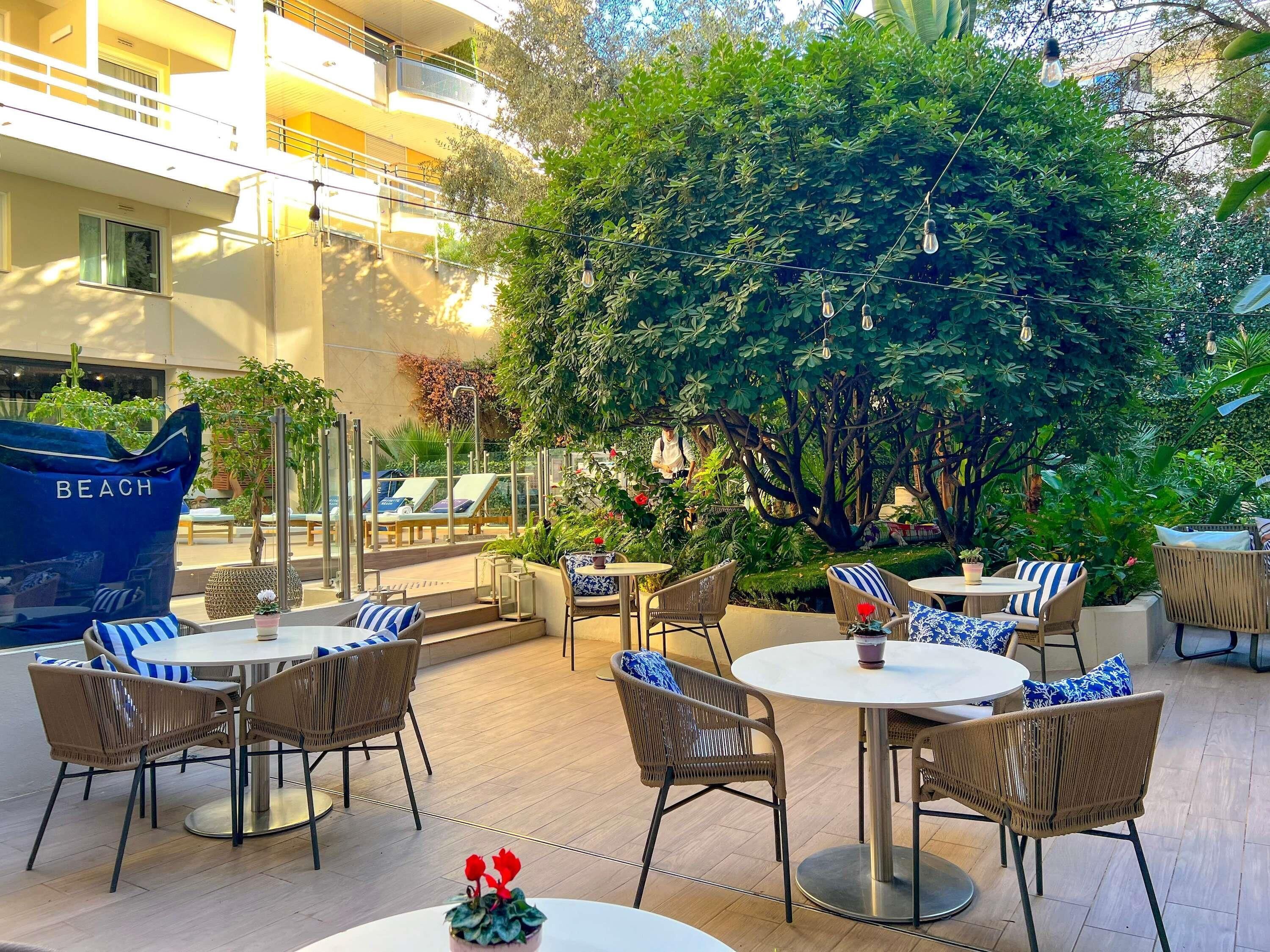 An outdoor seating area with wicker chairs and round tables, surrounded by lush greenery and a large tree.