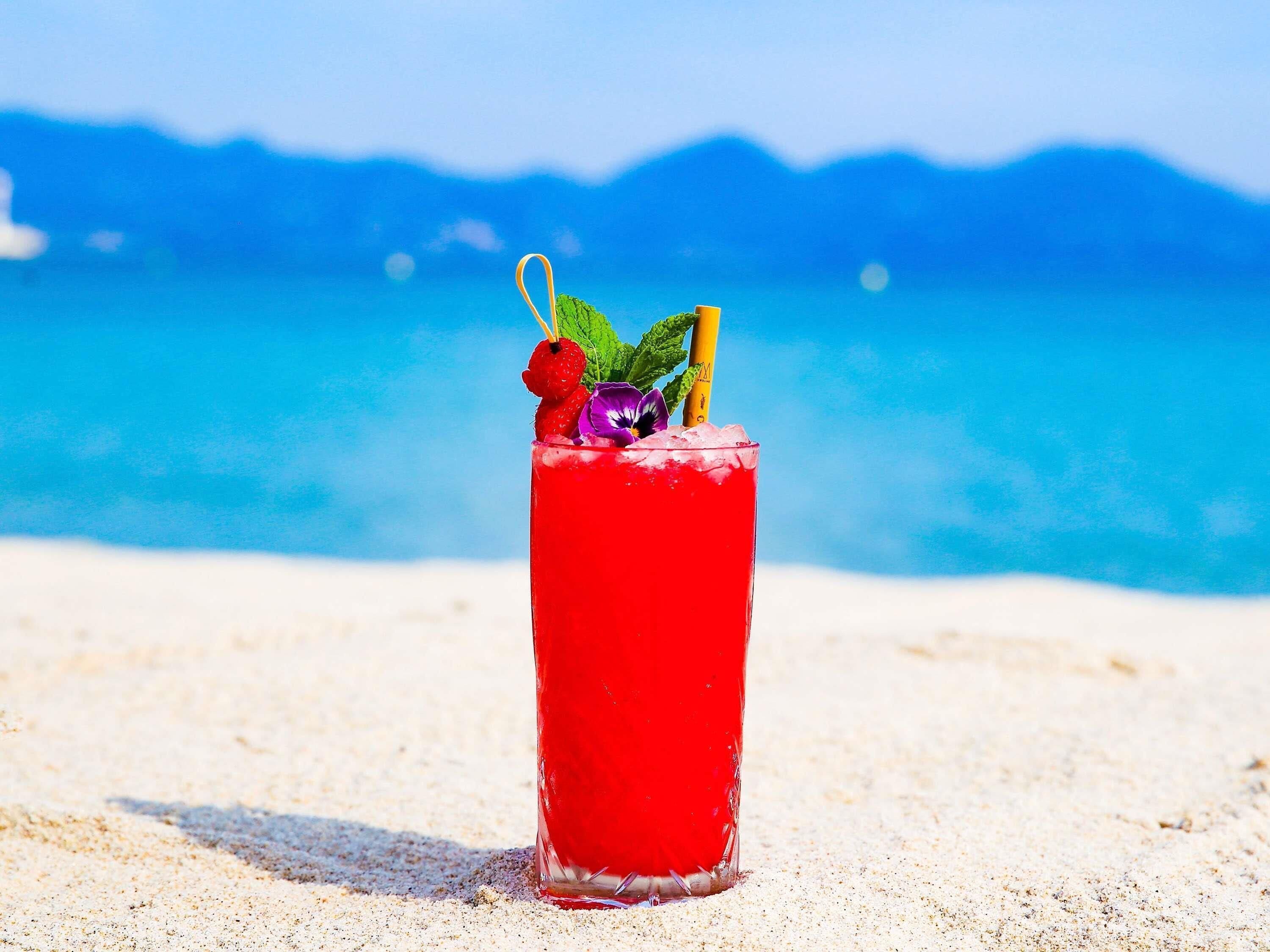 A tall glass of red beverage with strawberries and mint on a beach with a clear blue sky and ocean in the background.