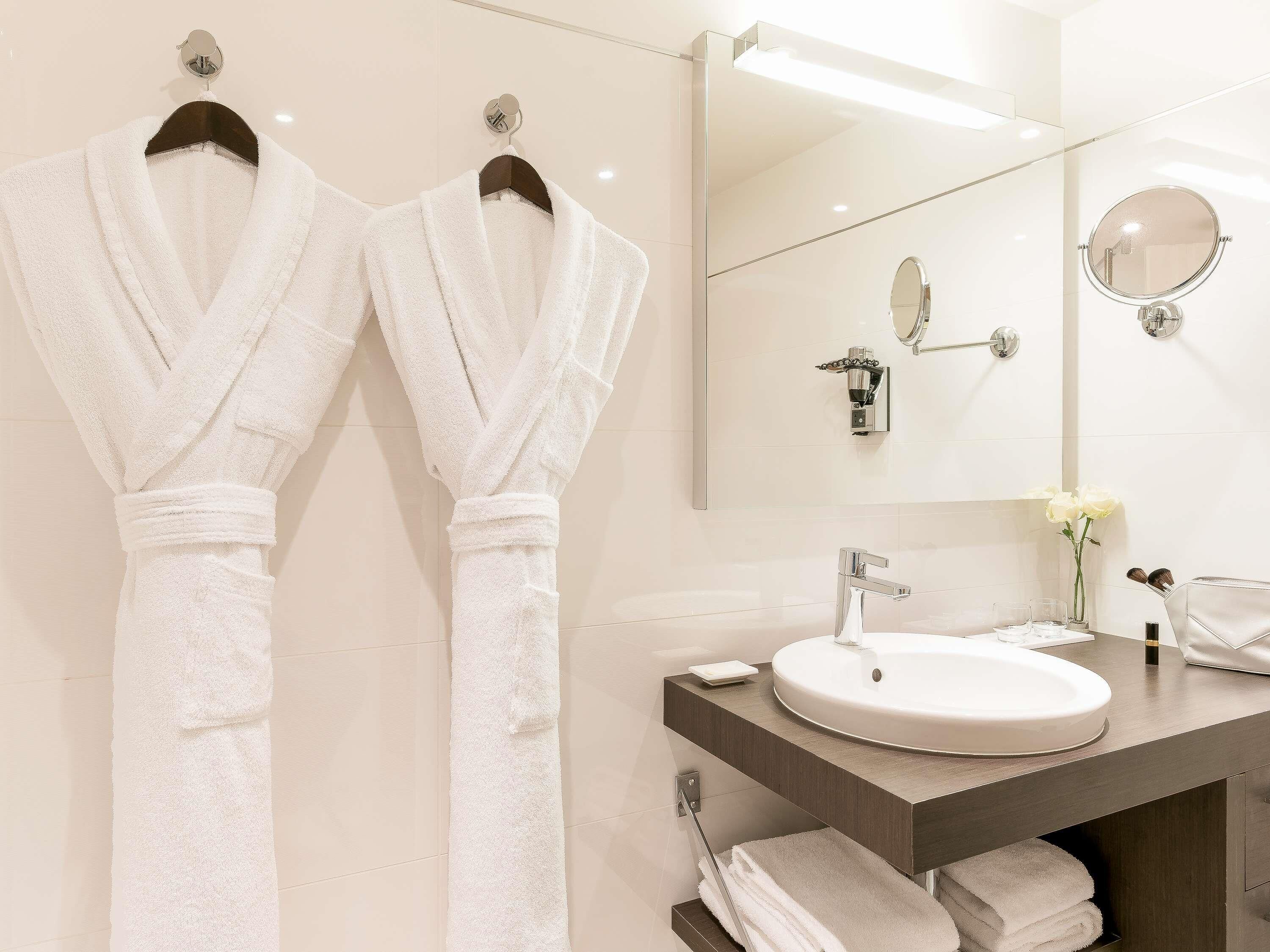 Une salle de bain avec deux serviettes blanches suspendues à des crochets, un miroir rond, un lavabo orné d’un vase de fleurs et un porte-serviettes.