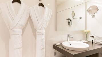 Une salle de bain avec deux serviettes blanches suspendues à des crochets, un miroir rond, un lavabo orné d’un vase de fleurs et un porte-serviettes.