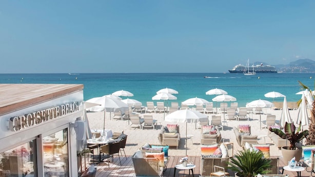 Een café aan het strand met terras, parasols en uitzicht op de zee en cruiseschepen.
