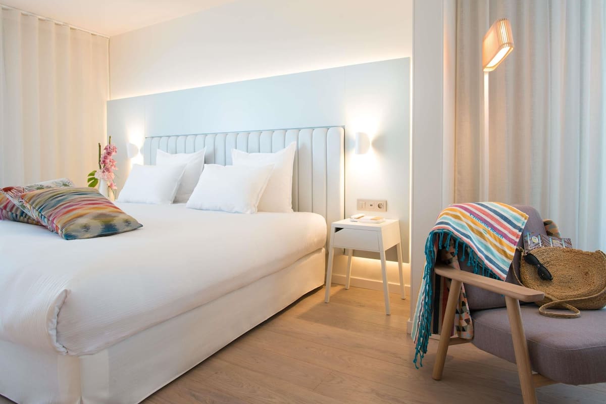 Ein Hotelzimmer mit Bett, Nachttisch, Lampe, Sessel und Blick ins Freie.