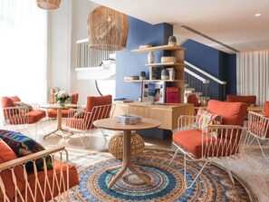A modern hotel lobby with a central round table, seating area with chairs, a bookshelf, and a staircase.