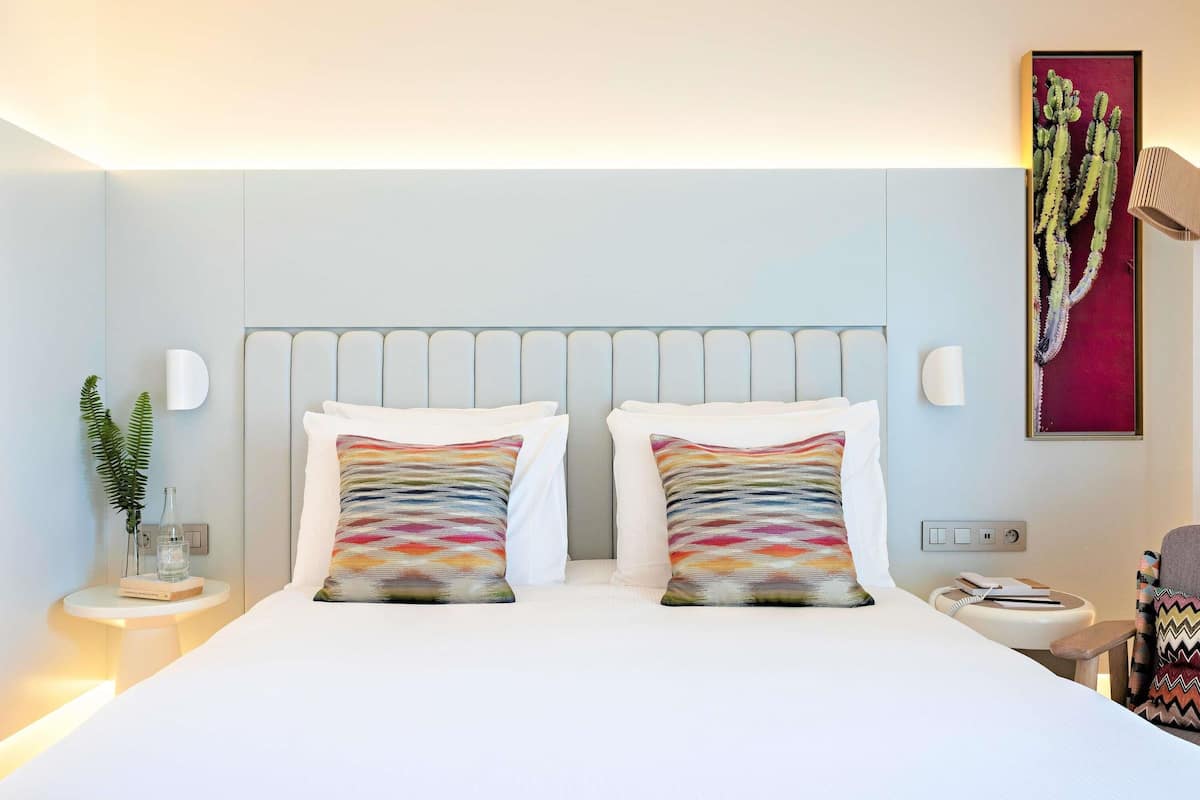 Ein modernes Hotelzimmer mit Bett, zwei Kissen, einem Nachttisch, einer Lampe, einem Wandbild und einer Pflanze.