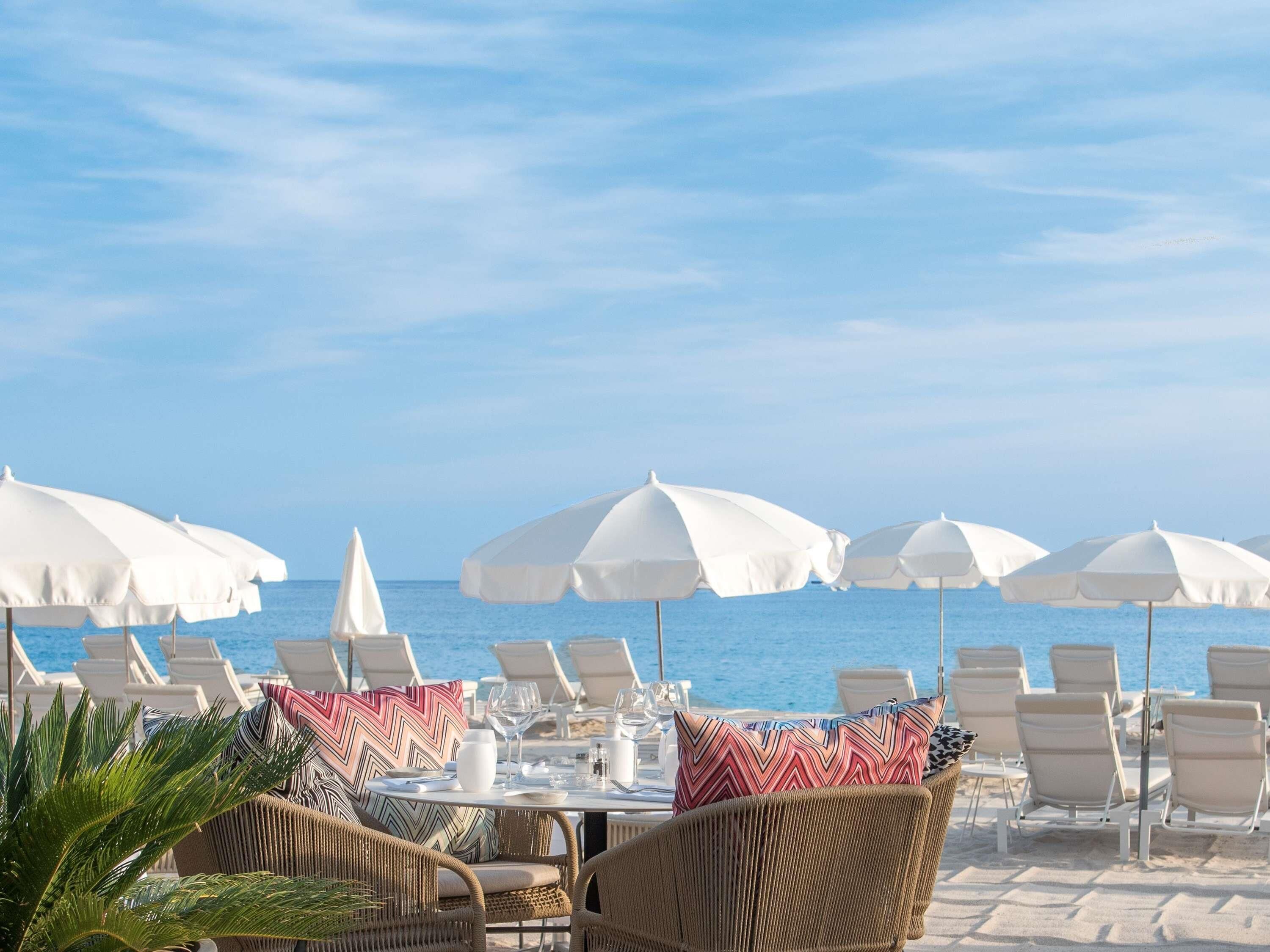 Un restaurante frente a la playa con terraza al aire libre, sombrillas blancas y vistas al océano.