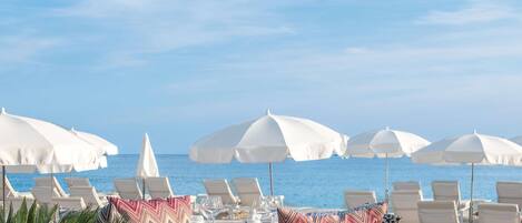 Un restaurante frente a la playa con terraza al aire libre, sombrillas blancas y vistas al océano.