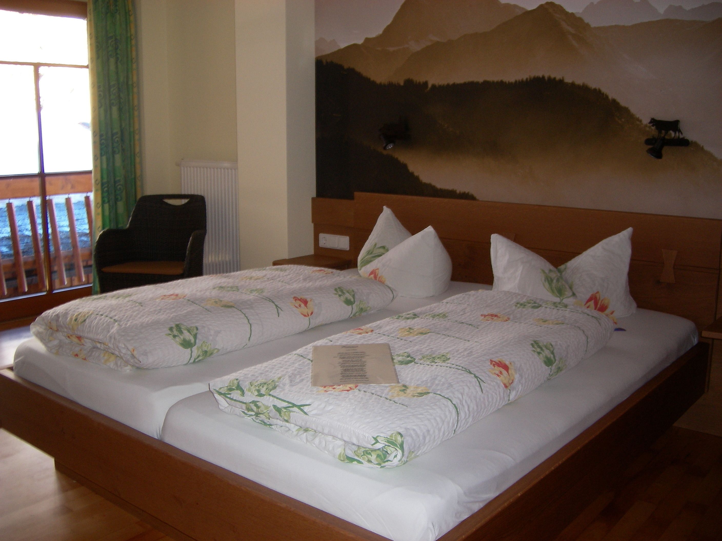 Cama doble con ropa de cama floral, una mesita de noche, una silla y un mural de un paisaje montañoso en la pared.