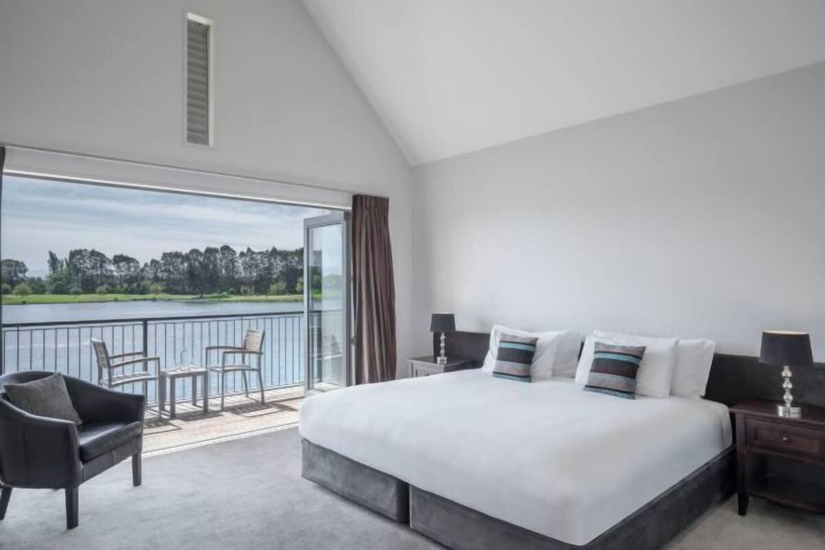 A bedroom with a large bed, a view of a lake, and a balcony with chairs.