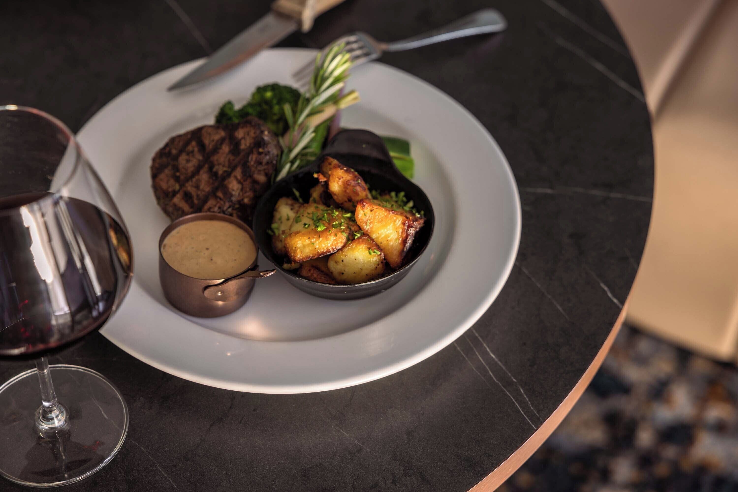 Un plato con bistec, papas asadas y vegetales de hoja verde, acompañado de una copa de vino tinto y un pequeño bol de salsa.