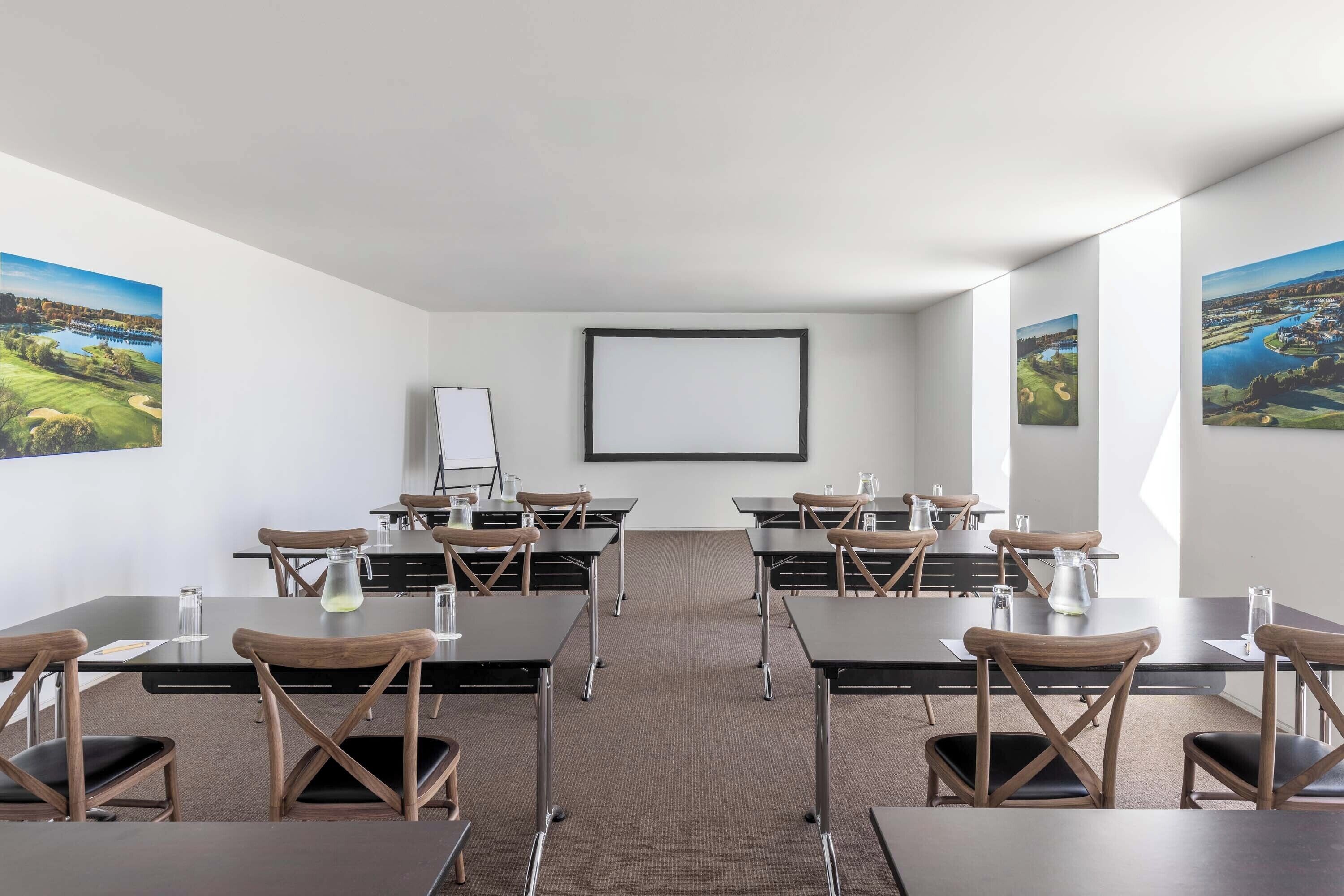 Ein Konferenzraum mit mehreren Tischen und Stühlen, einer Projektionsleinwand und gerahmten Landschaftsbildern an den Wänden.