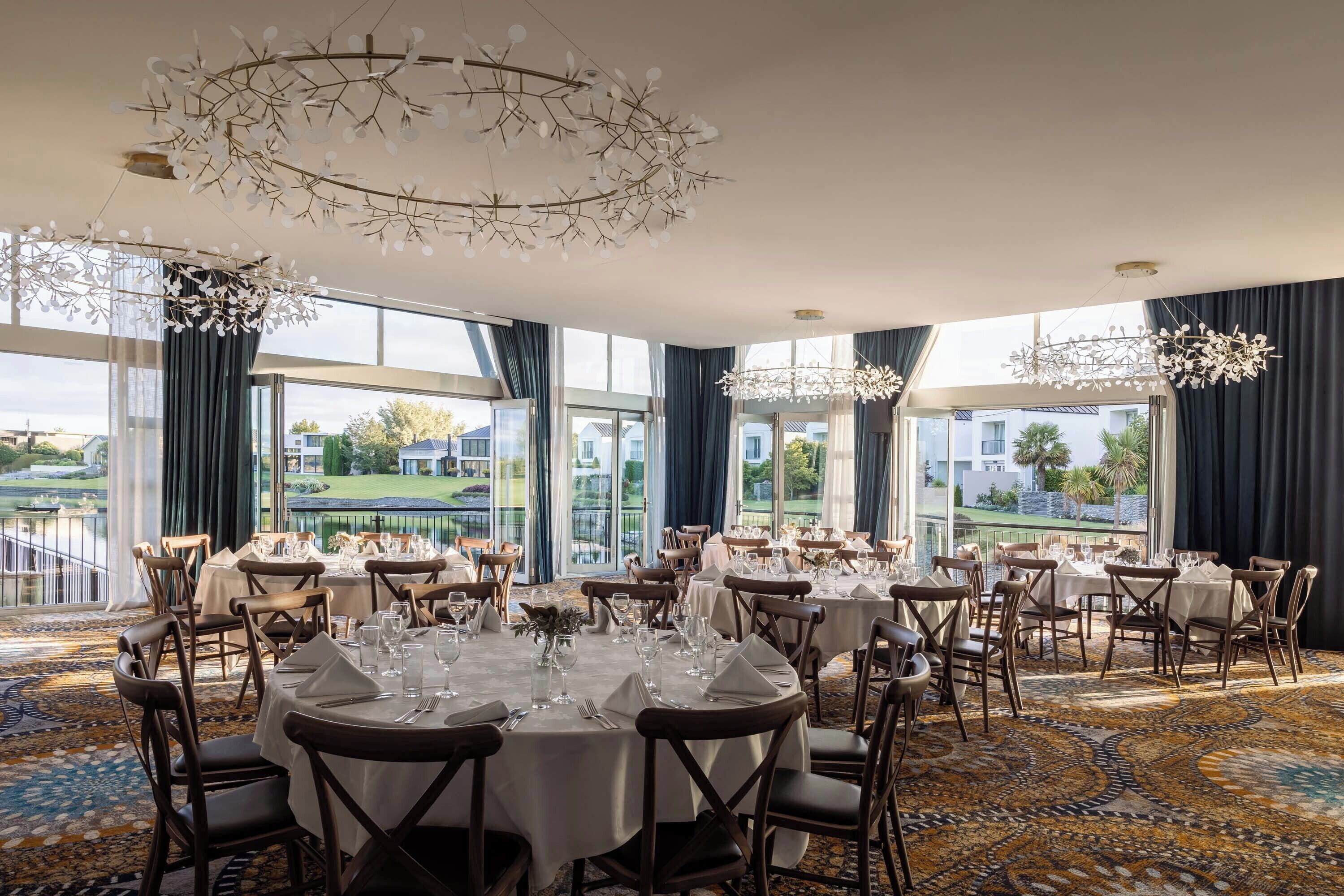 A dining area with round tables set for service, large windows offering a view of a golf course, and a chandelier with a leaf-like design.