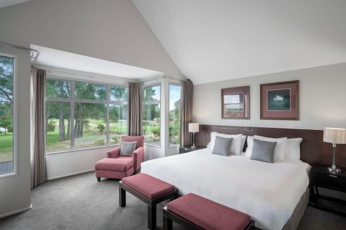 Habitación de hotel con una cama grande, un área de descanso con una silla y reposapiés, un escritorio con lámpara y vistas al exterior a través de amplias ventanas.