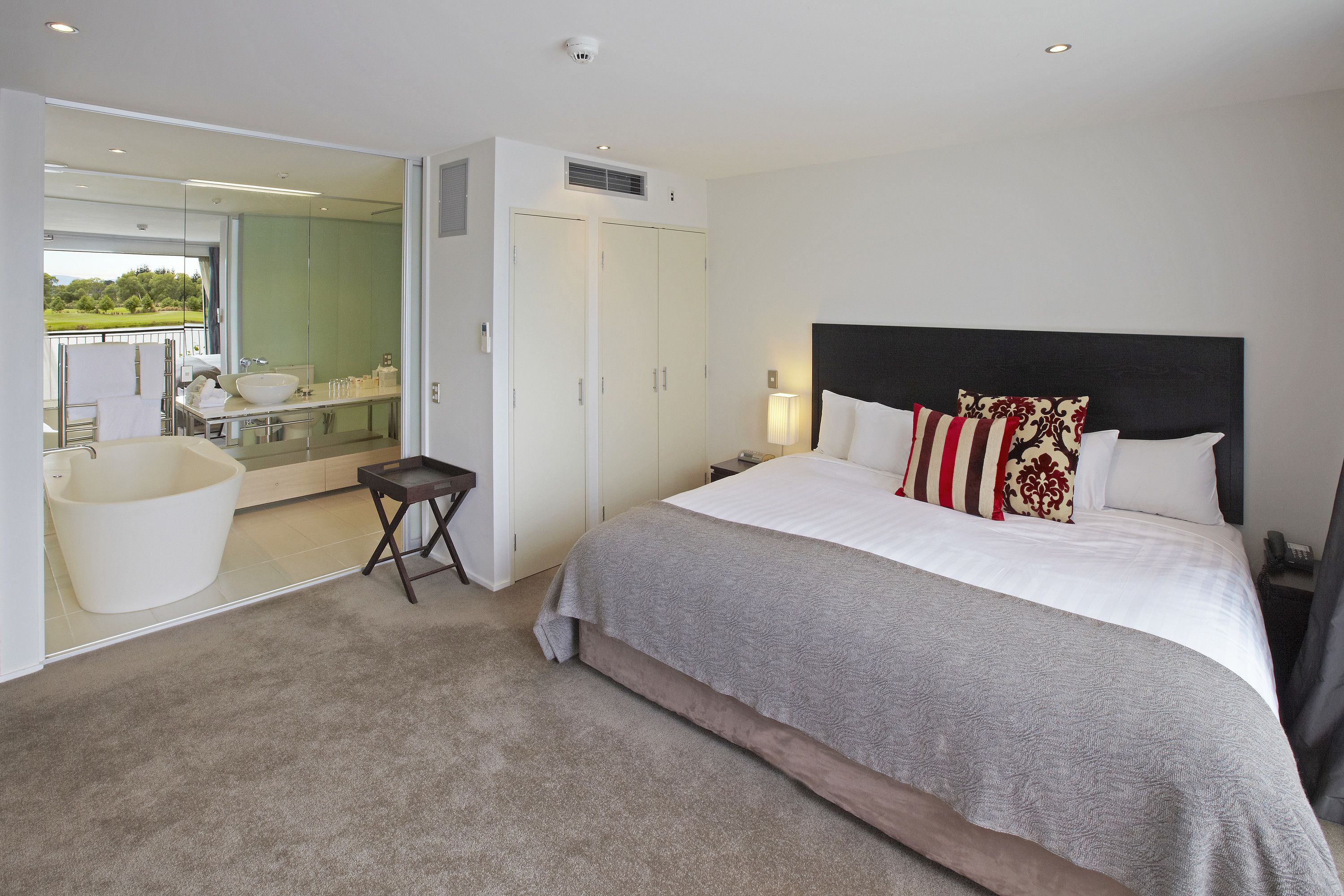 A modern bedroom with a large bed, a walk-in closet, a bathtub, and a view of a green landscape.