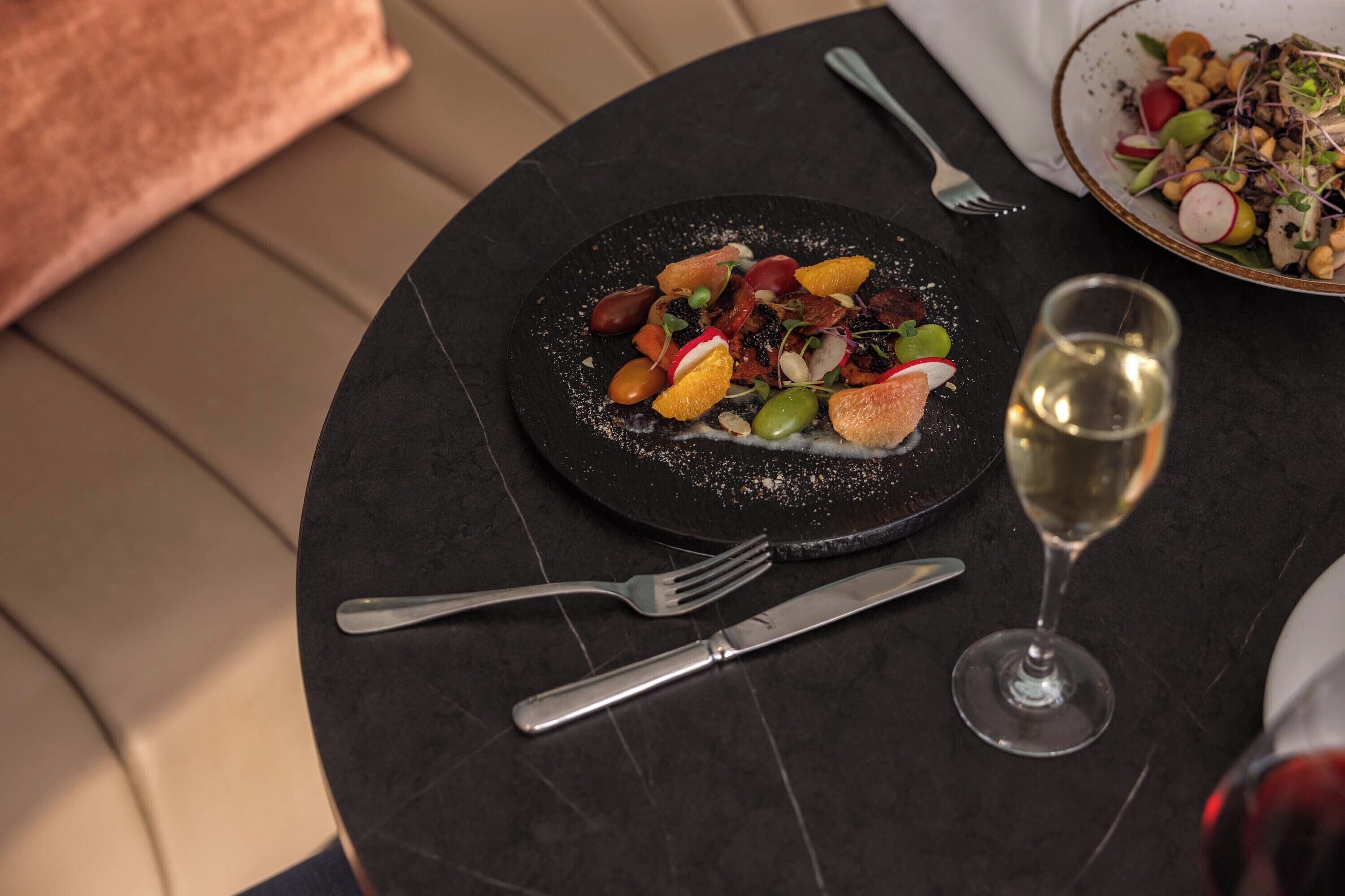 Une table de salle à manger ronde, avec une assiette noire contenant de la nourriture, un verre à vin et des couverts.