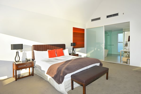 Un dormitorio de hotel moderno con una cama grande, una mesita de noche, un banco y un baño con paredes de vidrio.