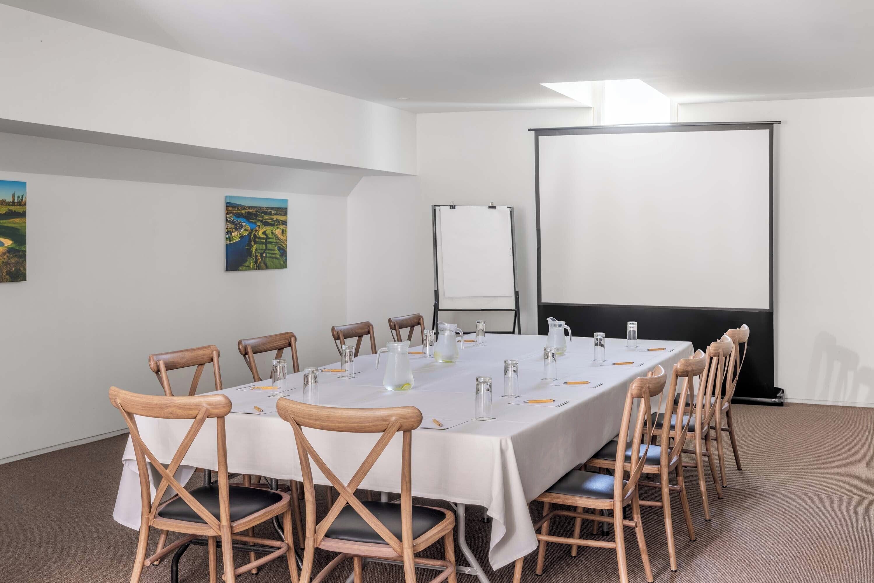 A conference room with a long table, wooden chairs, a projector screen, and two landscape paintings on the wall.