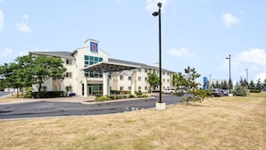 A modern hotel with a covered entrance, parking lot, and surrounding greenery. - Motel 6 Brampton, ON - Toronto (Brampton)