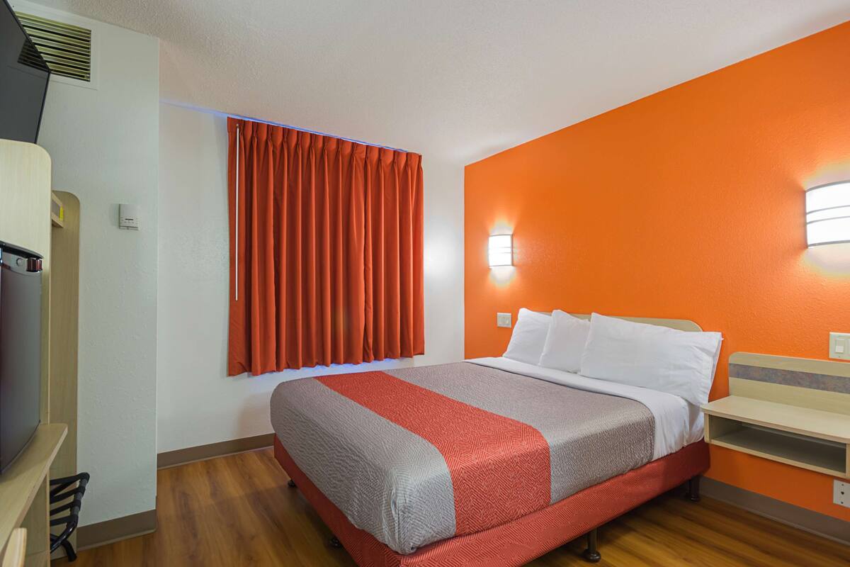 A hotel room with a bed, a TV, a window with curtains, and a wall-mounted light.